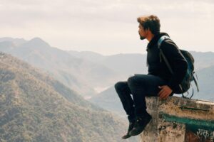 Photo of a young man sitting on the edge of a mountain, observing the beauty around him.